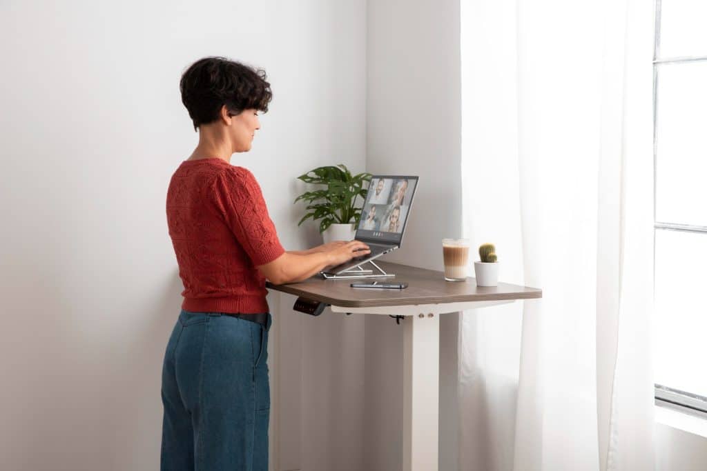Femme debout à son bureau en train de faire une visio. Elle a trouvé la hauteur de bureau idéale pour son bureau assis-debout.